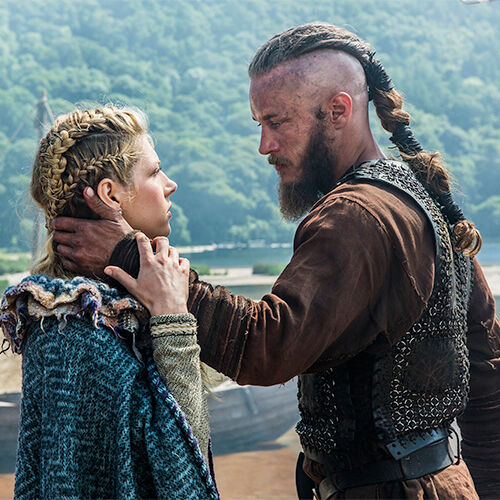 A viking woman named Lagartha stands face-to-face with a Viking man, who gently holds her face. Forested hills in the background.