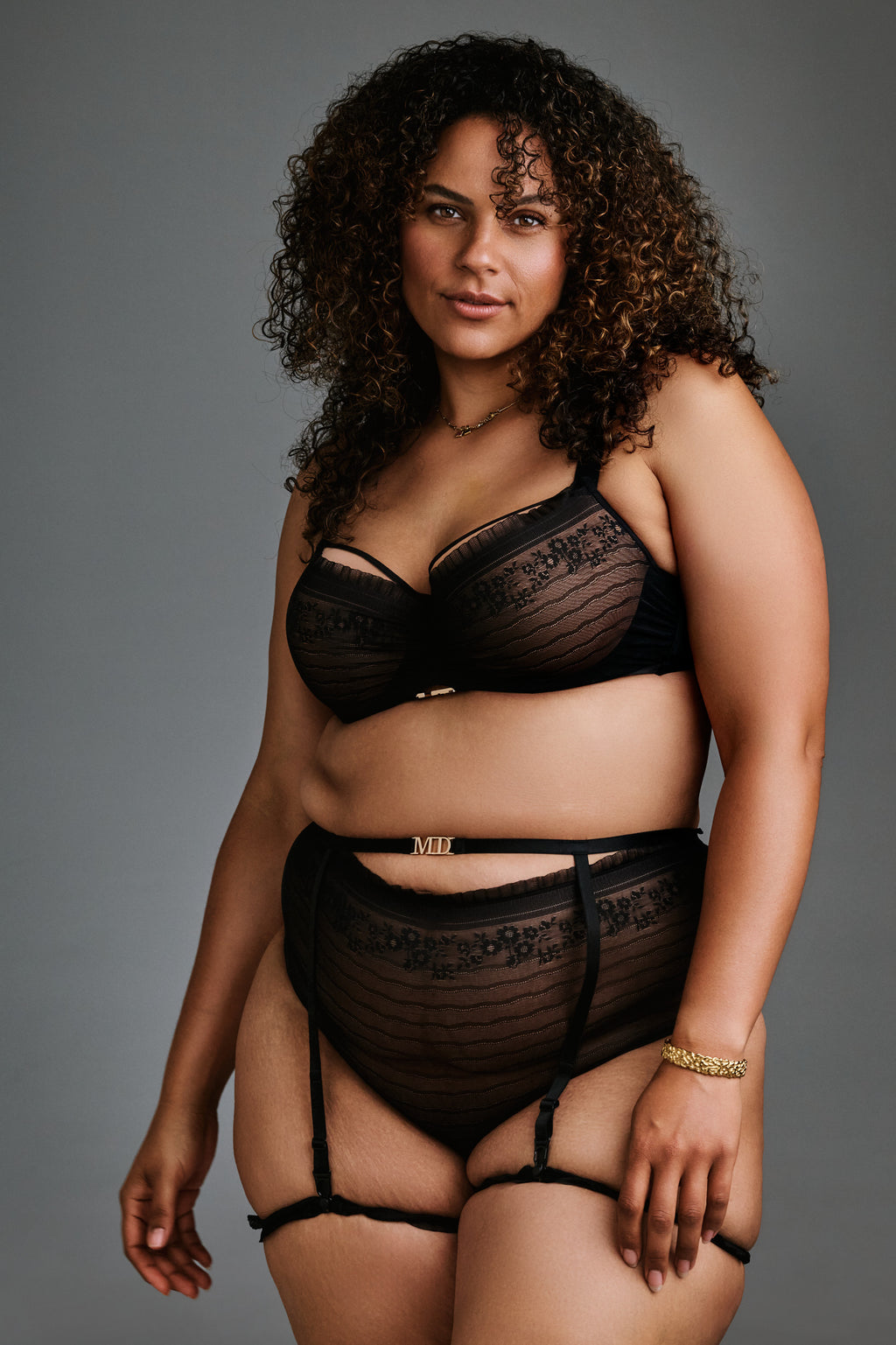 A plus-size model with curly brown hair wearing a black sheer lace lingerie set with garter straps, photographed against a solid gray studio background.